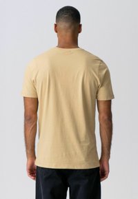 Beige pamut póló rövid ujjakkal, kerek nyakkivágással és egyenes szabással, hátulról nézve. Sima textúra, minimális részletek.