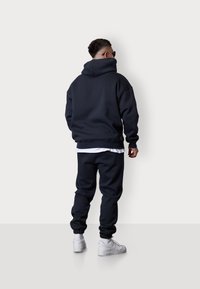 Marineblauer Kapuzenpullover und Jogginghose aus weichem Material, mit Kapuze, elastischen Bündchen und lockerer Passform, kombiniert mit weißen Turnschuhen.