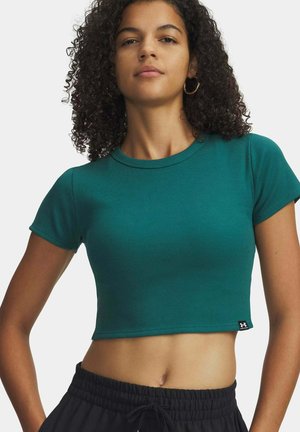 Vrouw met krullend haar, gekleed in een groene cropped geribbelde t-shirt en zwarte broek met elastische taille en trekkoord, tegen een effen achtergrond.