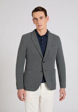 Man draagt een zwart-wit patroon colbert over een marineblauw poloshirt en witte broek tegen een effen grijze achtergrond.