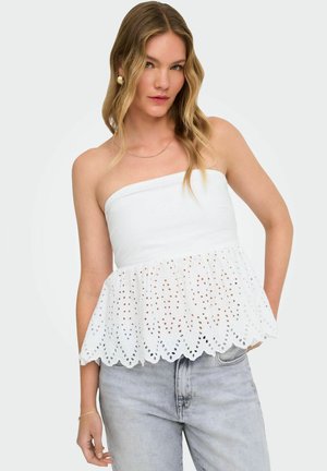 Kobieta z falującymi blond włosami, nosząca złote kolczyki, biały top bez ramiączek z ażurowej koronki i jasnoszare dżinsy, stojąca na jednolitym tle.