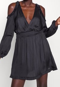 Svart satindress med djup V-ringning, kall axel-design, volangdetaljer, elastisk midja och en utsvängd kjol med volangkant.