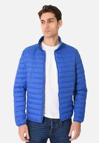 Giacca a vento blu con zip frontale, colletto alto, texture trapuntata e maniche lunghe. Indossata sopra una t-shirt bianca e un paio di jeans.