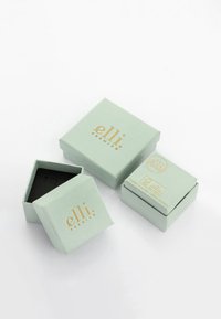Scatole di carta verde con accenti dorati. Una è quadrata e aperta, rivelando un interno nero. Le altre sono chiuse e presentano dettagli stampati.
