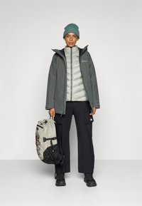 Grau wasserdichte Jacke über einer leichten, gepolsterten Innenschicht, kombiniert mit schwarzen Cargo-Hosen. Ein hellgrauer Rucksack mit dunklen Akzenten. Strickmütze in Türkis.