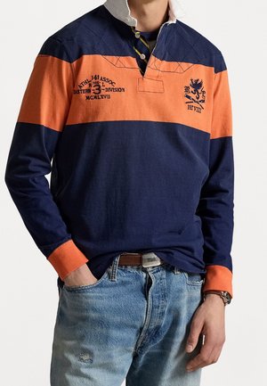 Mężczyzna ubrany w granatowo-pomarańczową koszulkę rugby z haftowanym tekstem i herbem, w połączeniu z jasnoniebieskimi dżinsami oraz zegarkiem na lewym nadgarstku.