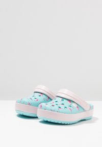 Kinderen clogs, lichtblauw met een roze flamingo patroon, gemaakt van rubberachtig materiaal. Voorzien van gaatjes voor ademend vermogen en een roze band.