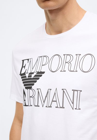 Balta kokvilnas T-krekls ar izteiksmīgu melnu tekstu "EMPORIO" un "ARMANI", stilizētu logotipu un vienkāršu apaļu kakla izgriezumu.