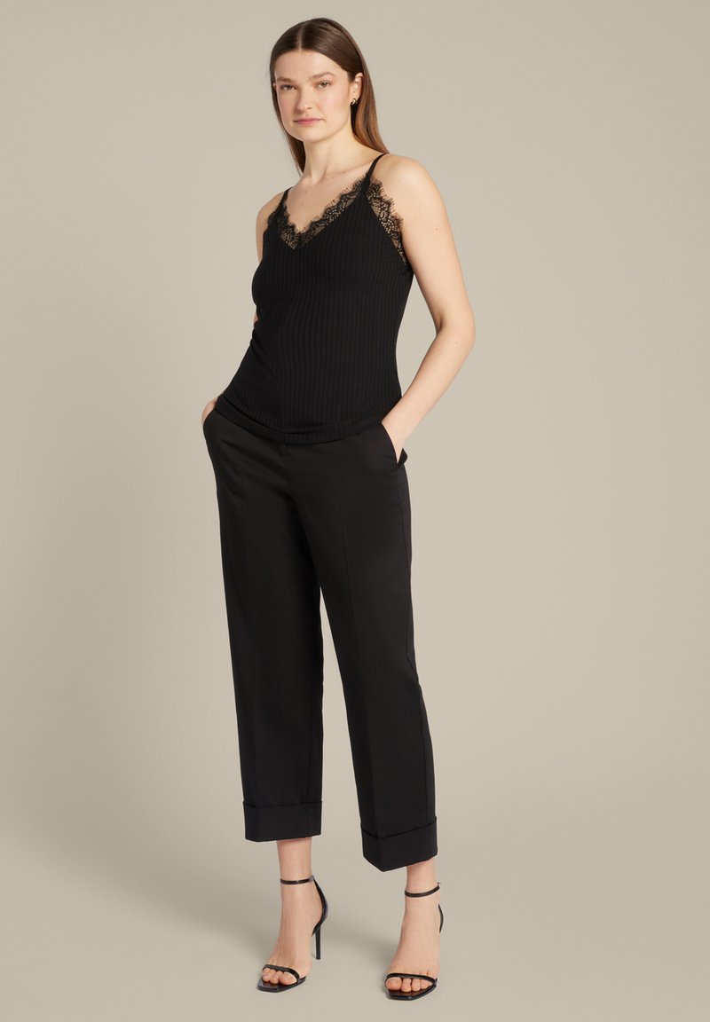 Zwarte geribbelde top met kantafwerking, gecombineerd met op maat gemaakte zwarte broek met omslagen. Model staat met handen in de zakken. Stilettohakken.