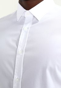 Camisa blanca de vestir con cuello clásico, con una textura suave y seis botones blancos a lo largo del frente.