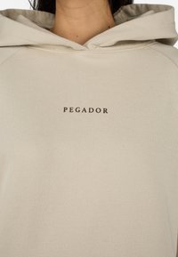 Cream-colored sweatshirt with a hood, featuring a dark brown "PEGADOR" logo in the center. Smooth texture and casual fit.