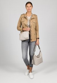 Veste kaki, haut beige clair, jean gris et baskets blanches. Deux sacs : un petit sac bandoulière et un grand sac à épaule beige avec détail de pompon.