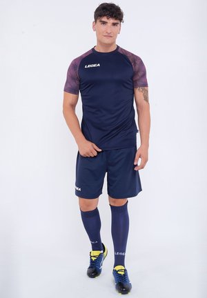 Giovane uomo in piedi che indossa abbigliamento sportivo blu navy con maniche viola a motivo, pantaloncini abbinati, calze lunghe e scarpini con dettagli gialli.