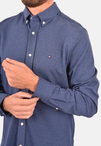 Camicia azzurra con bottoni realizzata in tessuto strutturato, con colletto e piccolo logo sul petto. I polsini sono arrotolati, mostrando i dettagli dei bottoni.