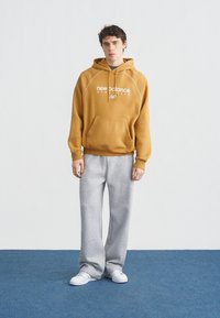 Νεαρός άνδρας στέκεται, φορώντας ώχρα κίτρινο φούτερ New Balance, γκρι φόρμα και λευκά αθλητικά παπούτσια σε μπλε χαλί με απλό φωτεινό φόντο.