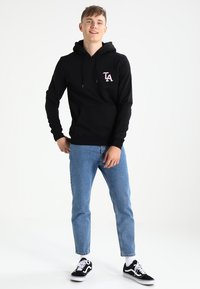 Zwarte hoodie met logo in het roze op de borst, kangoeroezak, capuchon met trekkoord, gecombineerd met lichtblauwe spijkerbroek en zwarte sneakers.