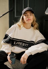 Jeune femme portant une casquette noire et un sweat-shirt de l'Université Liberty, assise sur des marches, tenant une tasse de café à emporter, regardant vers la caméra.