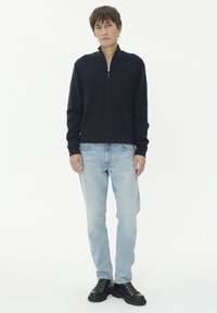 Tamsiai mėlynas užtrauktukas megztinis, ilgomis rankovėmis, pagamintas iš medvilnės mišinio, derinamas su šviesiai mėlynomis džinsomis ir juodais avalynės bateliais, stovintis prieš baltą foną.