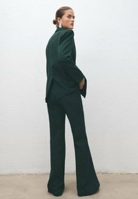 Tamnozeleni, krojeni blejzer s oštrim ramenima i proširenim hlačama, izrađen od čvrstog materijala. Istaknute srebrne naušnice i minimalistički dizajn.