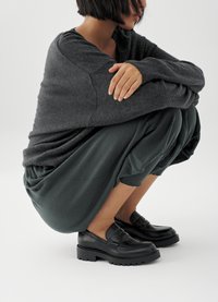 Svarte skinnloafers med en tykk såle, kombinert med en mørkegrå strikkegenser og løse, mørkegrønne klær, sittende i en hukende stilling.