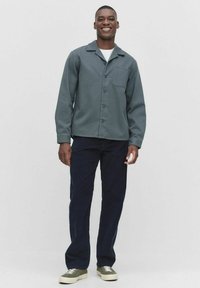 Grijze blouse met kraag en borstzak, gecombineerd met donkerblauwe broek en grijze sneakers. Gladde textuur, ontspannen pasvorm.