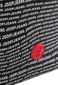 Czarna torba z tkaniny z powtarzającym się białym logo "JOOP!JEANS", ze prominentnym czerwonym okrągłym akcentem na dole.