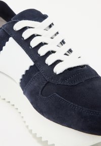 Baskets en suède bleu marine avec des accents en cuir blanc, dotées de lacets blancs et d'un design en zigzag texturé. Semelle en caoutchouc blanc épais.