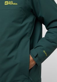 Veste imperméable vert foncé avec une finition texturée. Les caractéristiques incluent des poignets ajustables avec Velcro et des éléments de marque jaunes.