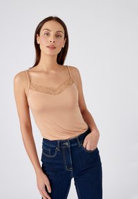 Camisola beige chiaro con pizzo lungo il collo, spalline sottili e tessuto morbido. Abbinata a jeans a vita alta blu scuro.
