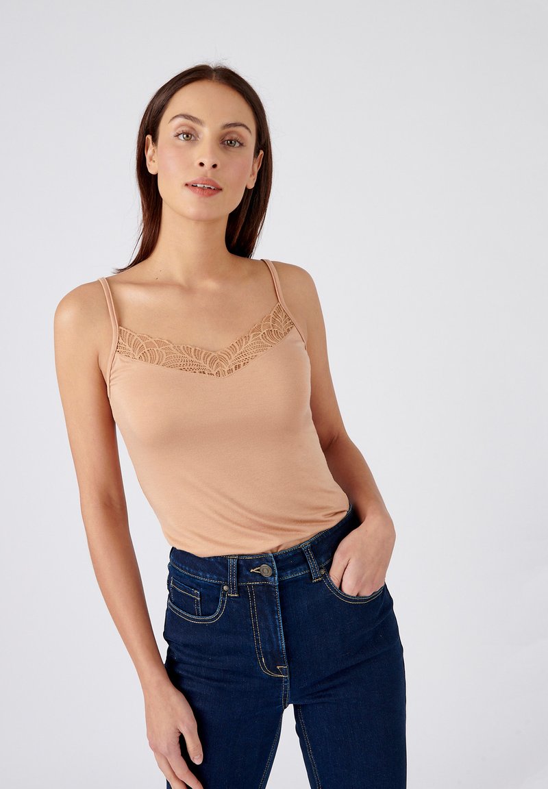 Camisola beige chiaro con pizzo lungo il collo, spalline sottili e tessuto morbido. Abbinata a jeans a vita alta blu scuro.