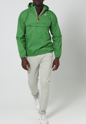 Grüne Pulloverjacke mit Kapuze, ausgestattet mit einer Reißverschlusstasche, einem kontrastierenden gestreiften Kragen und elastischen Bündchen. Kombiniert mit hellen Hosen und Turnschuhen.