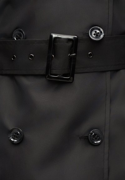 Τμήμα μαύρου παλτού τύπου trench coat που δείχνει τέσσερα μαρμαρωτά κουμπιά και μια φαρδιά ζώνη με ορθογώνιο μαύρο αγκράφα και μεταλλικές τρύπες.