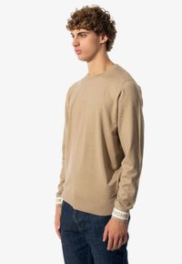 Suéter de punto de manga larga beige con cuello redondo, que presenta puños acanalados blancos con la impresión "RICH", combinado con jeans de mezclilla oscura.