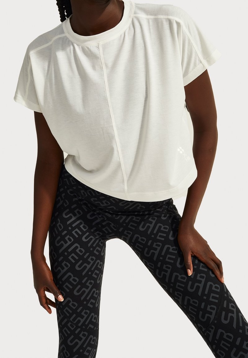 White short-sleeve athletic top with rounded neckline. Paired with black leggings featuring a prominent repeating logo pattern.