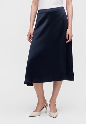 Un mannequin porte une jupe midi bleu marine foncé, un haut blanc et des escarpins beiges à bout pointu, sur un fond uni.