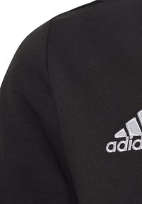 Black fabric garment with ribbed texture, showcasing a white embroidered adidas logo on the left chest area.
