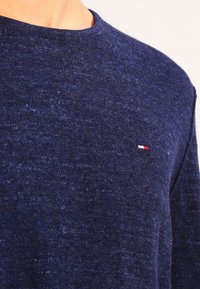 Marinblå stickad tröja med rund halsringning och liten rektangulär röd, vit och blå logotyp på vänster bröst.