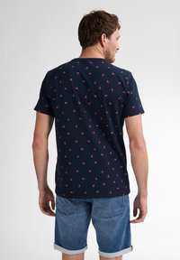 Tummansininen t-paita, jossa on pieniä punaisia kuvioita, lyhyet hihat ja pyöreä kaula-aukko, valmistettu pehmeästä puuvillasekoitteesta. Päällä farkkushortsit.