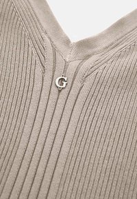 Gros plan sur un tissu beige en tricot côtelé avec un col en V et une petite lettre métallique "G" cousue près du centre.