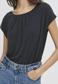 Schwarzes kurzärmliges Oberteil mit plissiertem Ausschnitt, aus weichem Stoff, getragen zu hochgeschnittenen Jeans aus Denim mit einer verblassten blauen Textur.