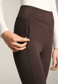 Brązowe legginsy wykonane z elastycznego materiału, posiadające boczną kieszeń na zamek i wysoki pas. Gładka tekstura z minimalnymi detalami szwów.
