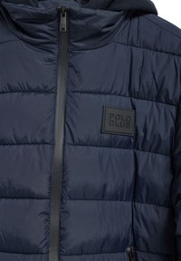 Mörkblå puffjacka med horisontell sömnad, framtida dragkedjeförslutning, och en rektangulär svart logotyp med texten "POLO CLUB" på bröstet.