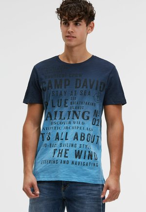 Junger Mann mit lockigem Haar trägt ein blaues Gradient-T-Shirt mit nautischem Schriftzug und blaue Jeans, steht vor einem schlichten Hintergrund.