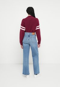 Bordó pulóver fehér csíkokkal az ujjain, világoskék bőszárú farmerrel párosítva. A pulóver laza szabású és bordázott textúrájú.