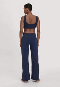 Vrouw die blootvoets staat, met haar rug naar ons toe, gekleed in een marineblauwe mouwloze crop top en wijdvallende broek tegen een effen lichte achtergrond.