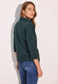 Felpa verde scuro con colletto, maniche a tre quarti e orlo dritto, abbinate a jeans blu con tasche posteriori.