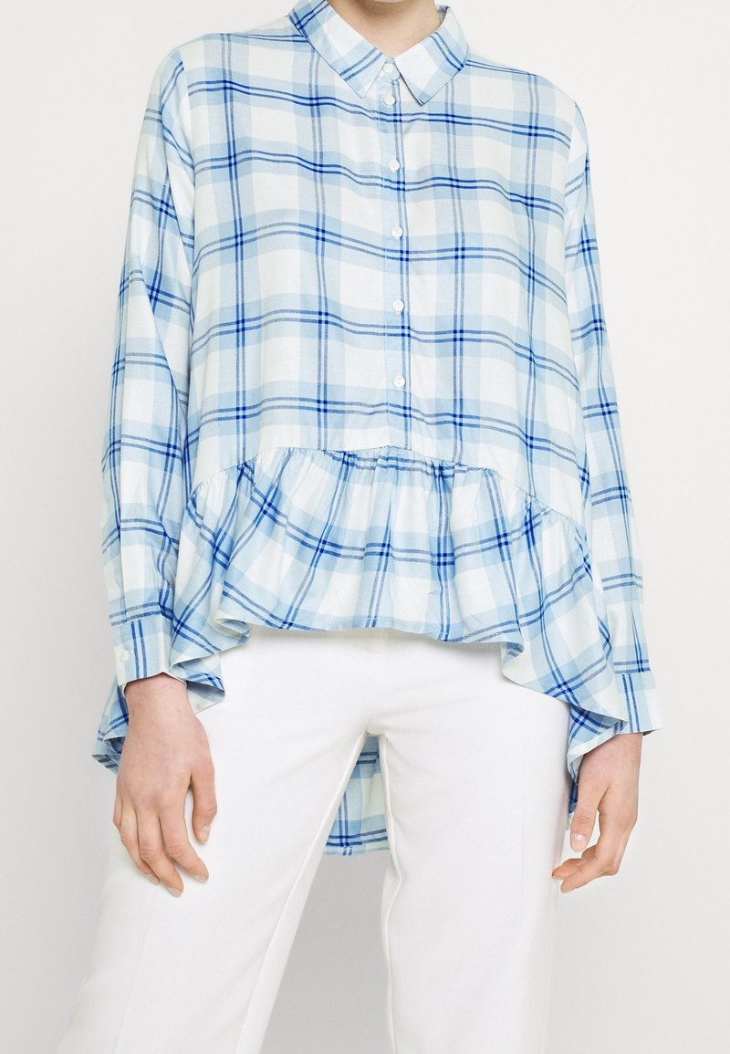Femme portant une chemise à manches longues à carreaux bleu et blanc avec un ourlet volanté, associée à un pantalon blanc sur un fond uni.