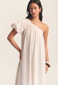 Abito asimmetrico a una spalla in tessuto beige chiaro con scollatura arricciata e manica a sbuffo; modella che indossa un bracciale su un polso.