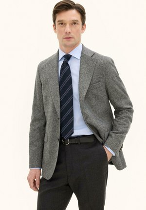 Mann trägt grauen Wollblazer, hellblaues Hemd, gestreifte marineblaue Krawatte, schwarzen Gürtel und dunkle Hose, steht vor einfachem hellem Hintergrund.