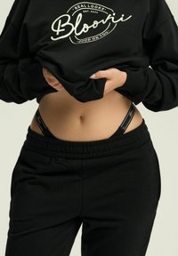 Czarna bluza z logo, w połączeniu z czarnymi joggerami. Widoczny czarny pas, na którym znajdują się drobne białe detale brandingowe.